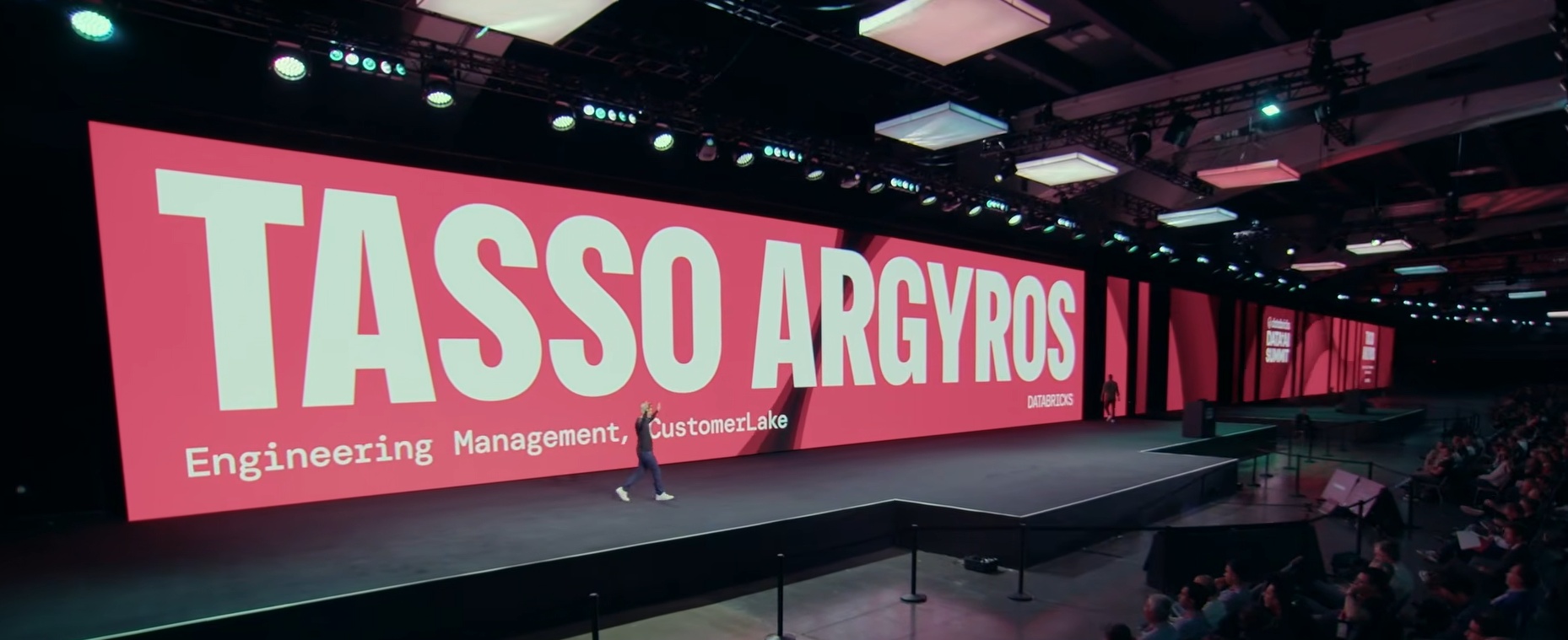 Tasso Argyros walks across the Data and AI Summit 2026 stage beneath a screen displaying his name
