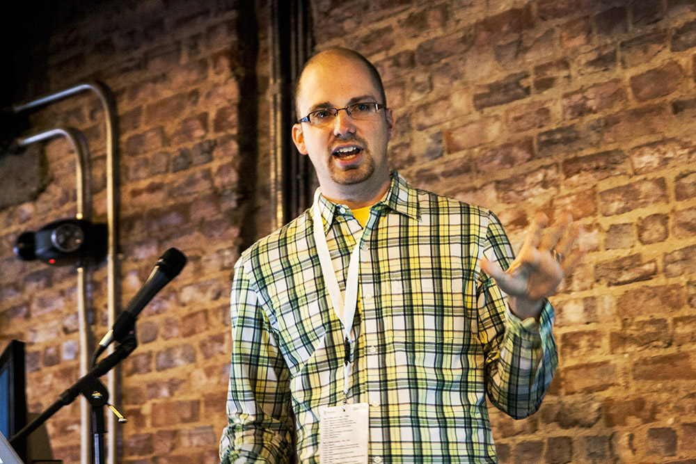 Taylor Otwell speaking into a microphone at Laracon DC in 2013, wearing a checked shirt in front of a brick wall