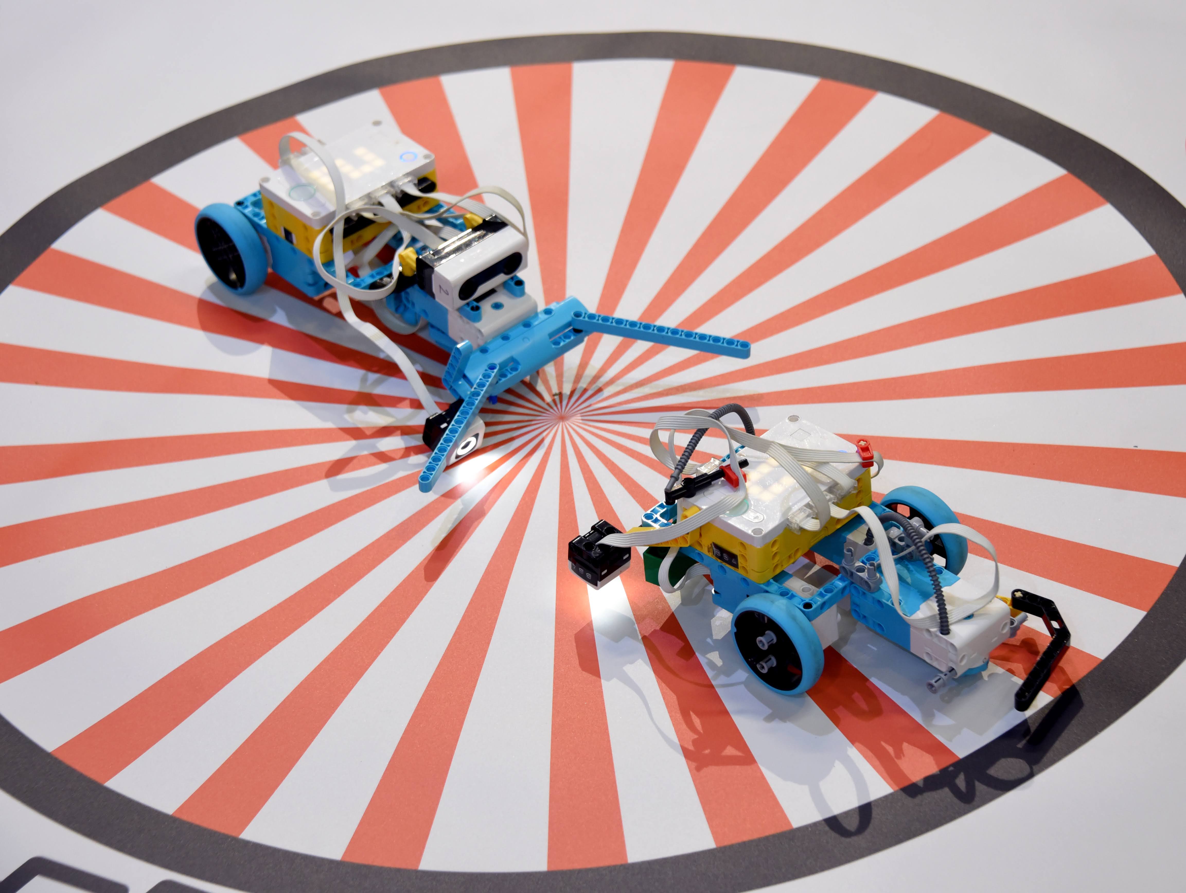 Two small wheeled robots facing each other on an orange and white circular arena