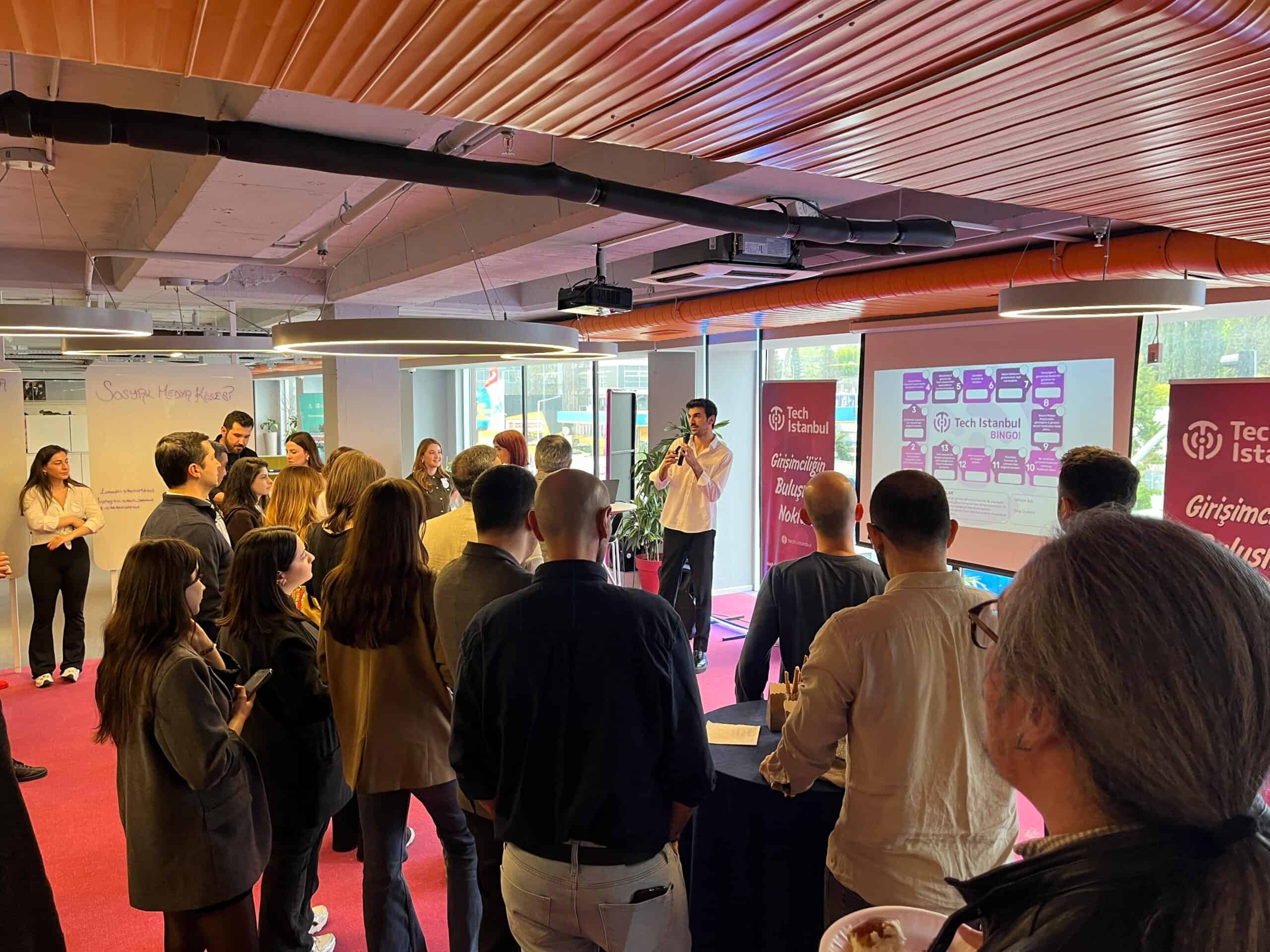 People gather around a speaker at Tech Istanbul’s Şişli location, with an entrepreneurship bingo slide projected behind him.