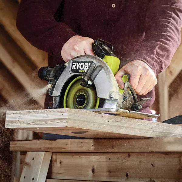 A person using a green RYOBI ONE+ circular saw to cut timber on a workbench