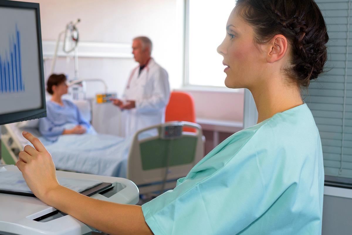 A clinician works at a hospital computer while a patient and doctor are visible in the room