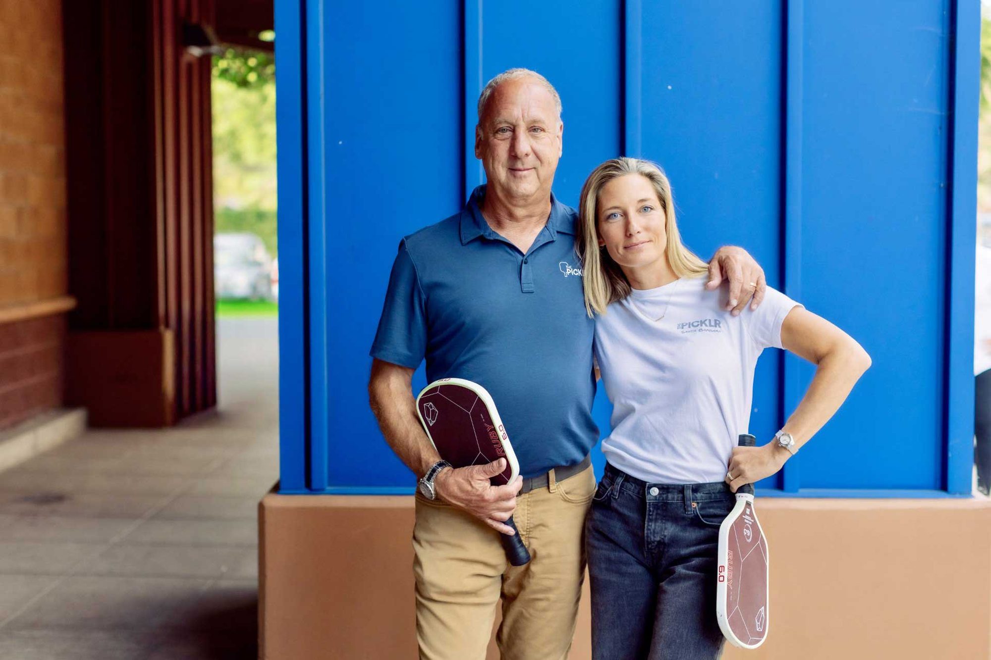Ted and Trish Guggenheim holding pickleball paddles in front of a blue wall