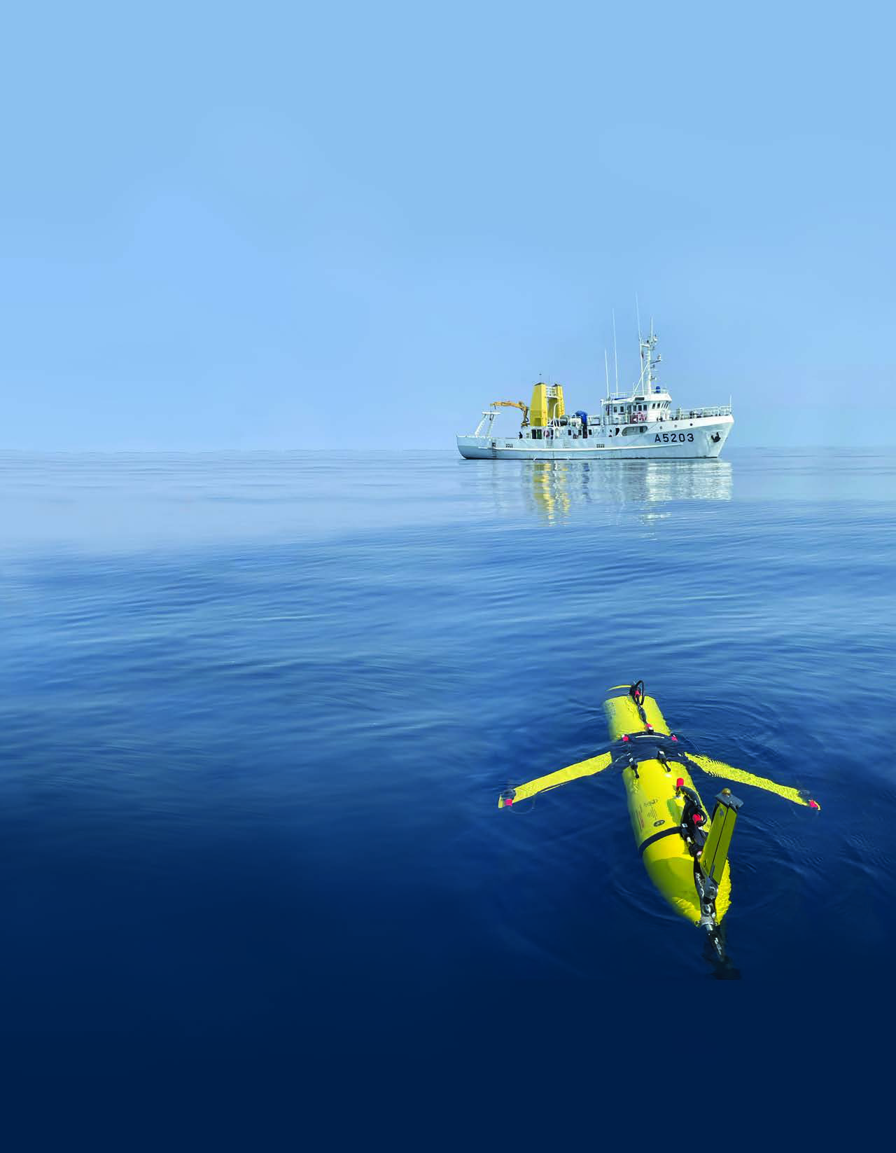 A yellow Slocum underwater glider floats in the foreground with a research vessel behind it