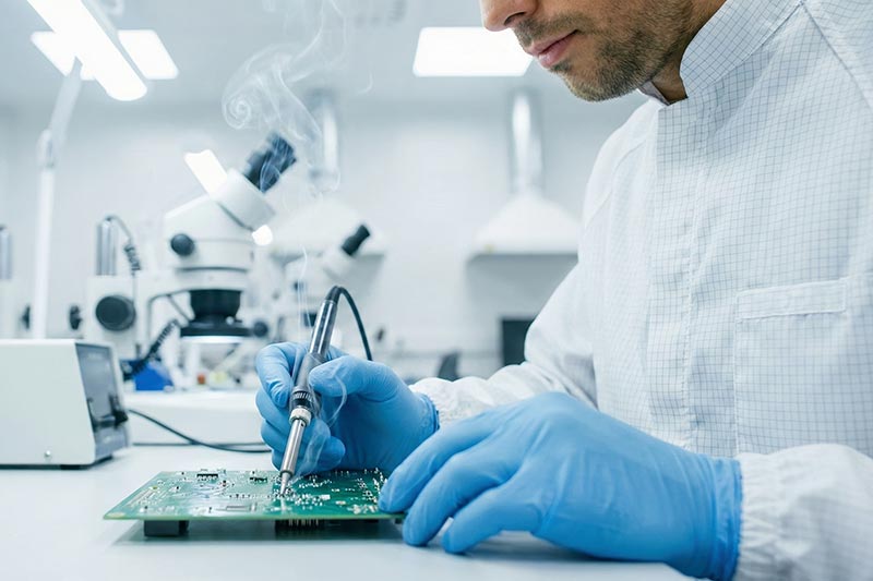 An electronics technician solders a circuit board beside laboratory inspection equipment
