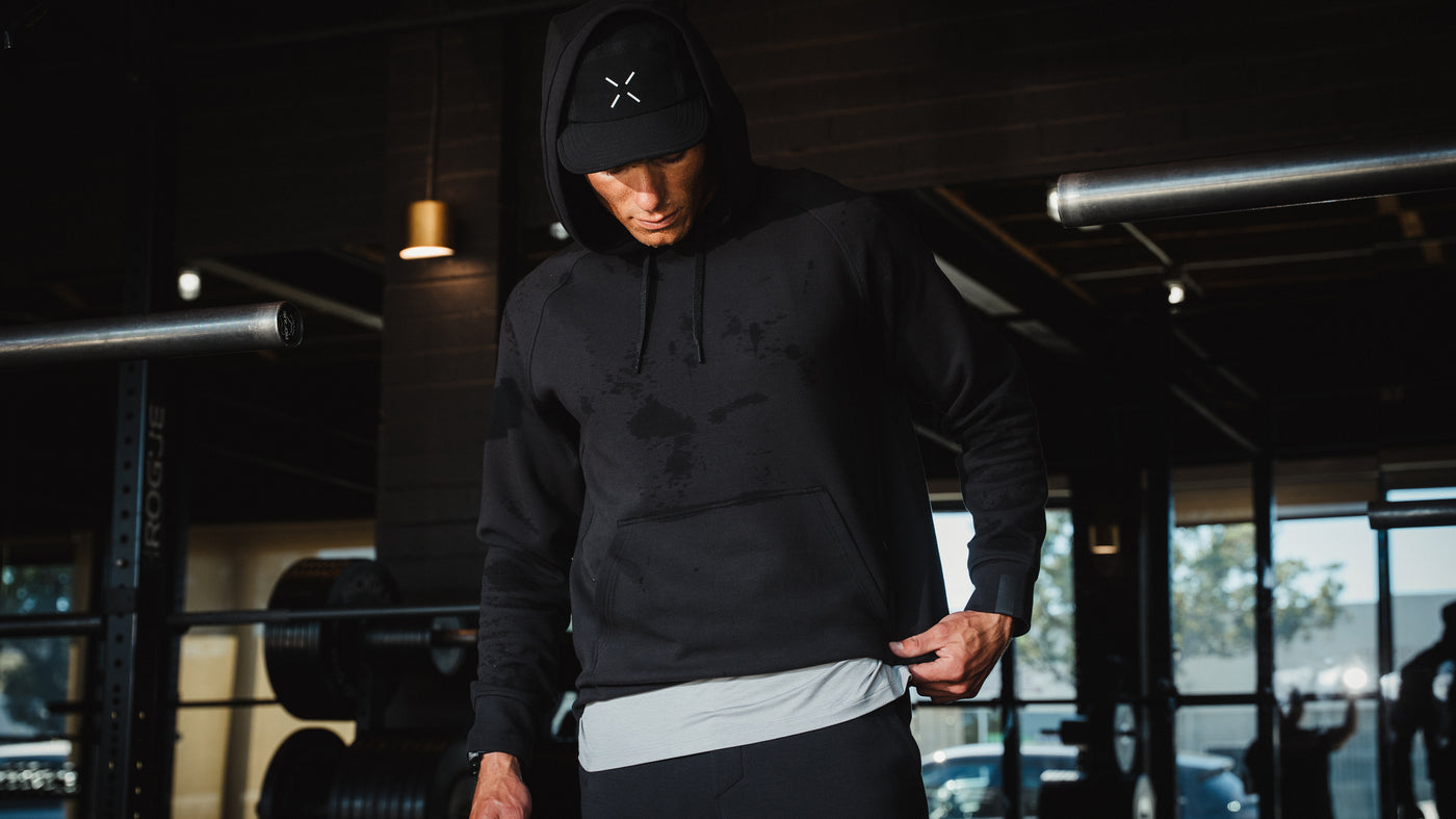 An athlete in a black Ten Thousand hoodie and cap beside gym equipment