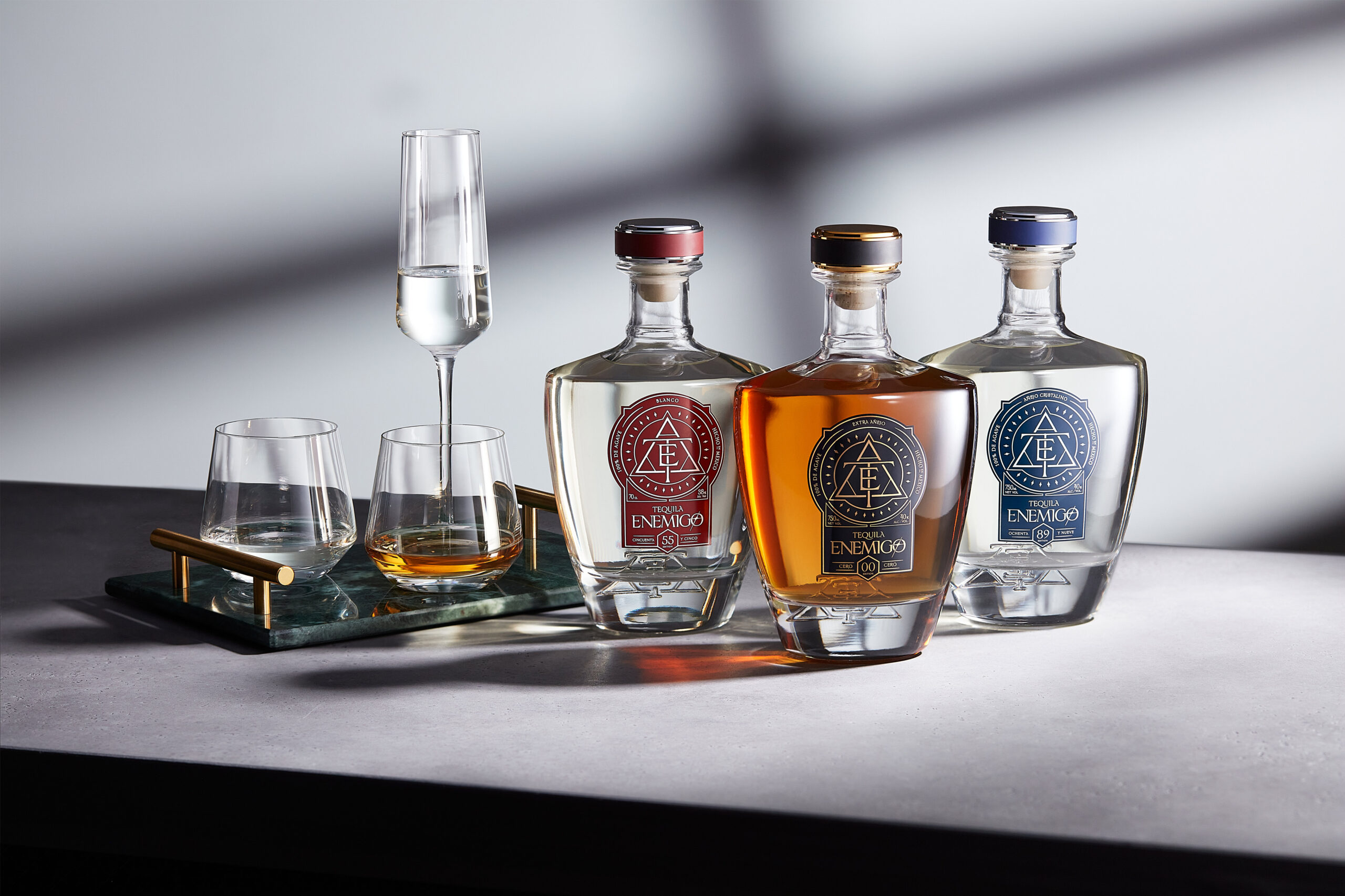 Bottles of Enemigo 55 Blanco, 00 Extra Añejo and 89 Añejo Cristalino arranged with tasting glasses