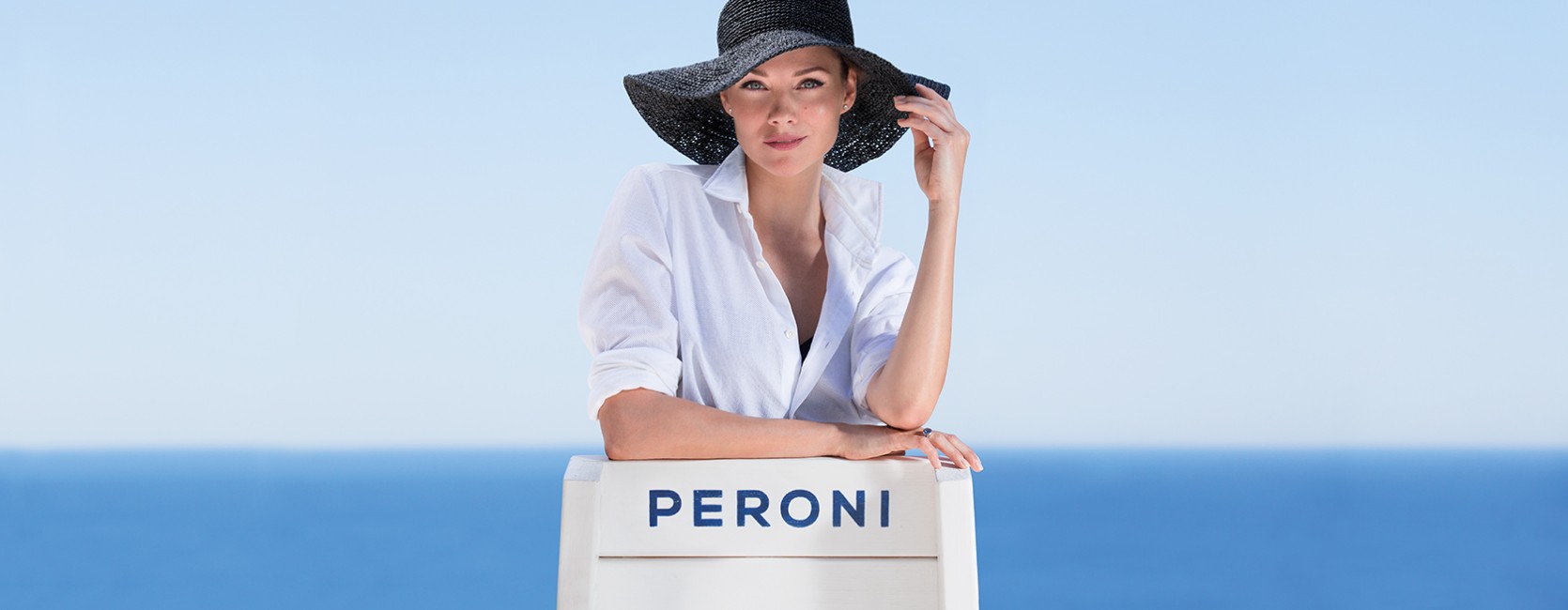 Peroni Vita al Mare campaign photograph showing a woman in a black hat beside a branded chair and the sea