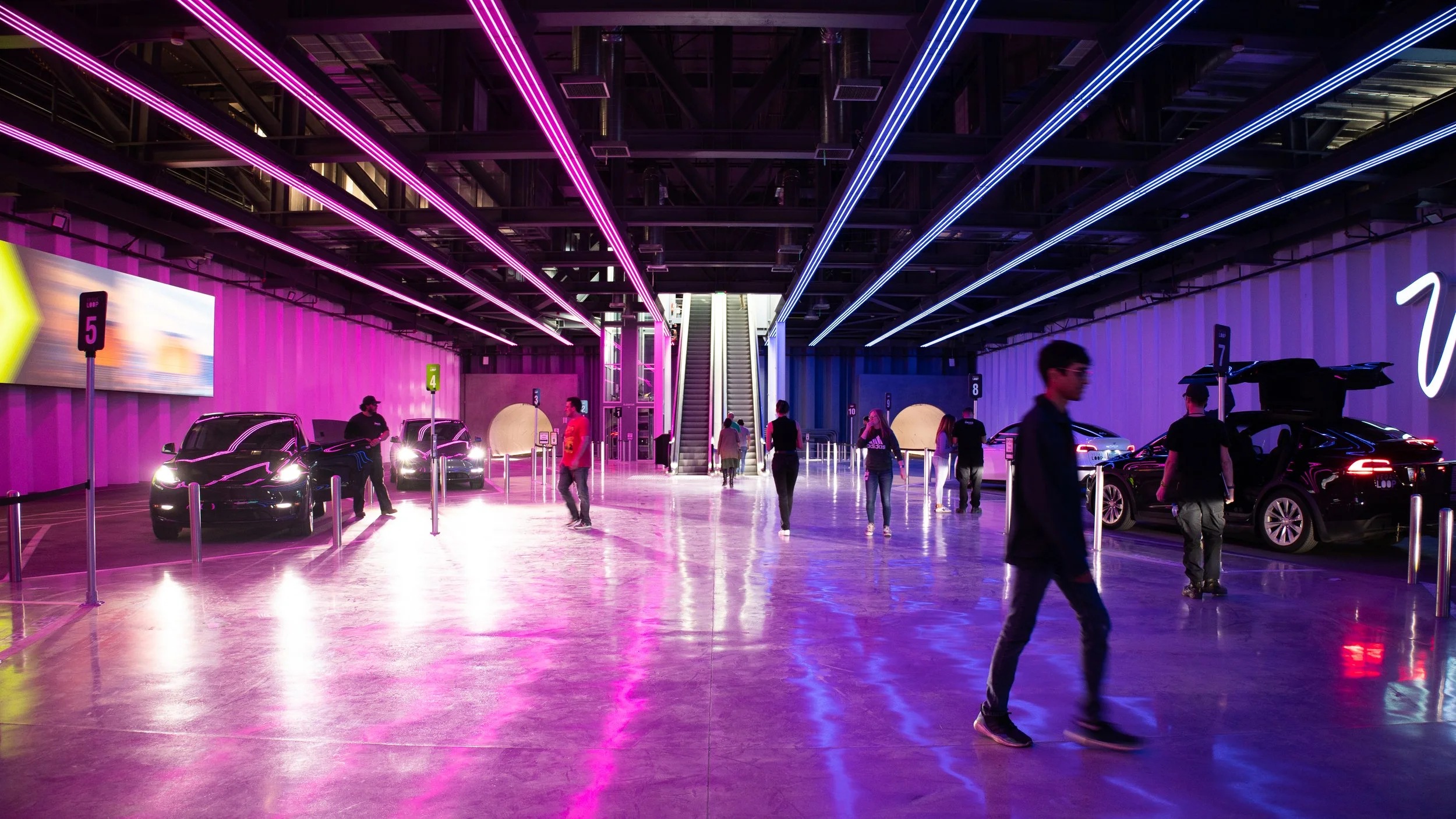 Tesla vehicles and passengers beneath pink and blue lights at LVCC Central Station