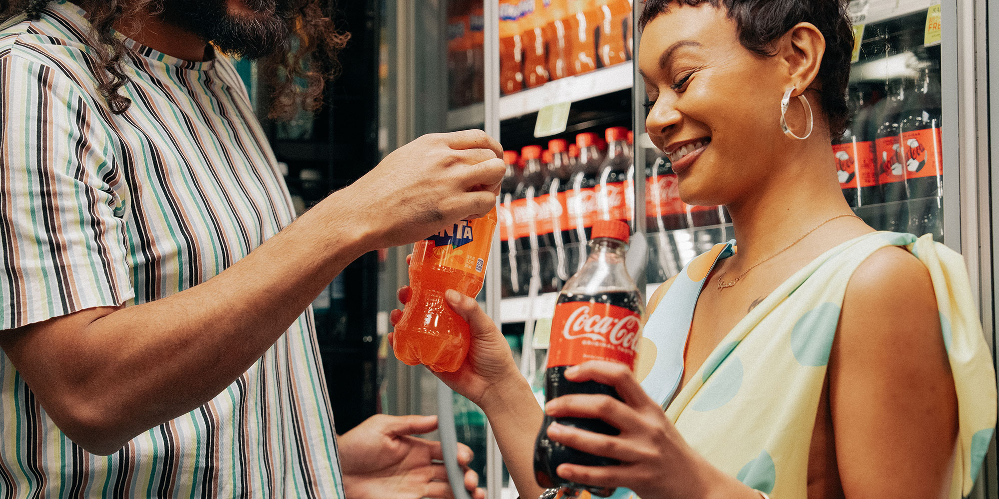 Two shoppers holding Coca-Cola and Fanta bottles in front of a drinks refrigerator