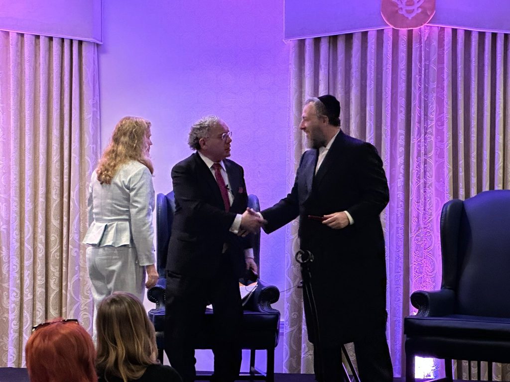 Ezra Friedlander shaking hands with a speaker at a public event while another guest looks on