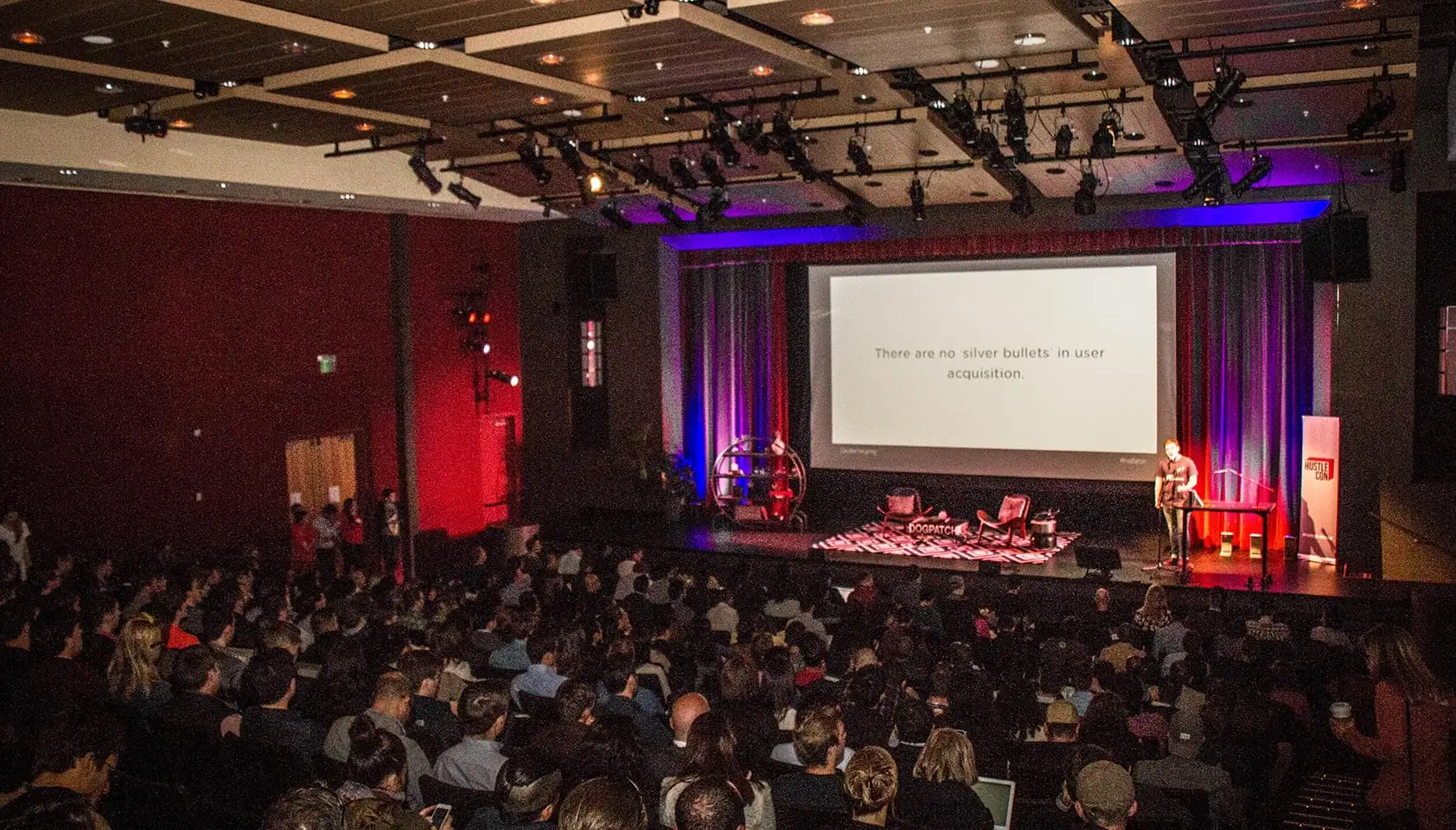 An audience at Hustle Con watching a speaker beneath a slide about user acquisition