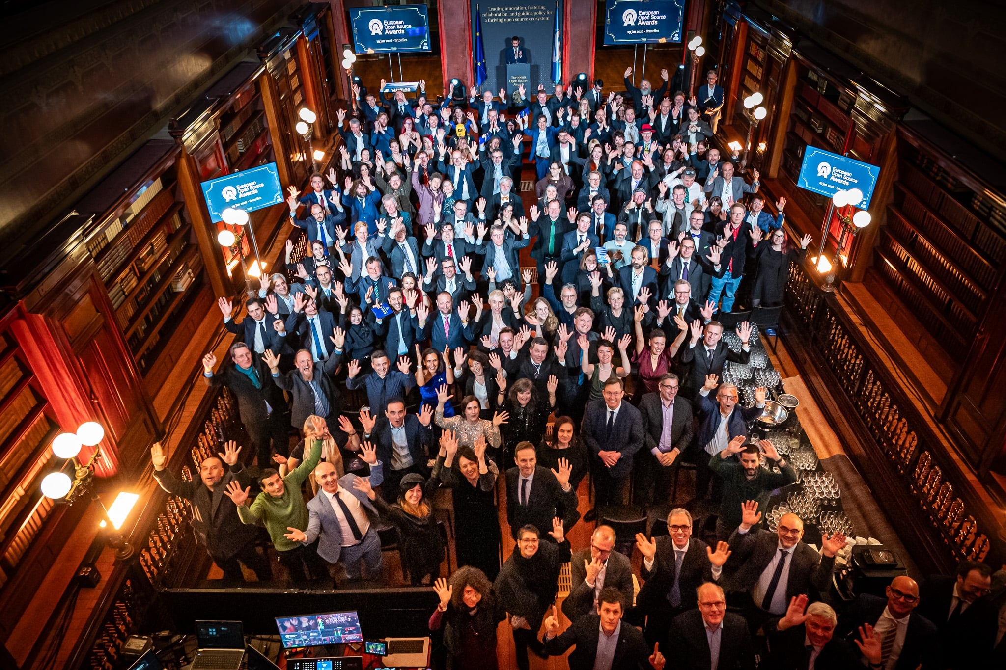 Participants wave to the camera at a European Open Source Awards gathering in a wood-paneled hall