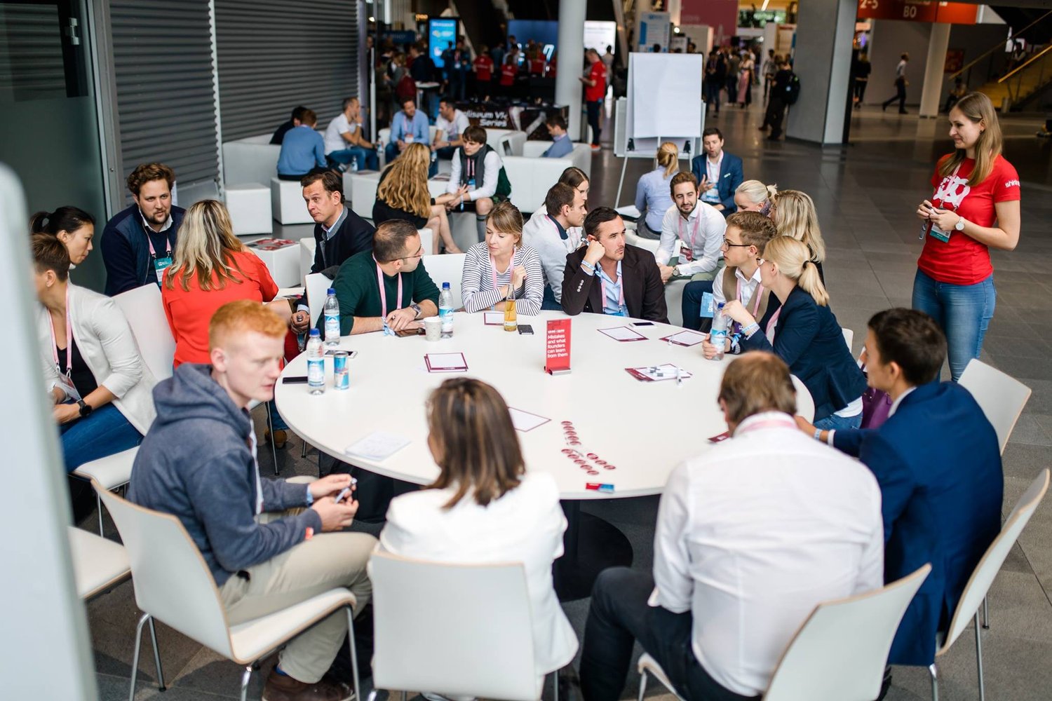 Attendees talking around a large roundtable with red-shirted facilitators nearby