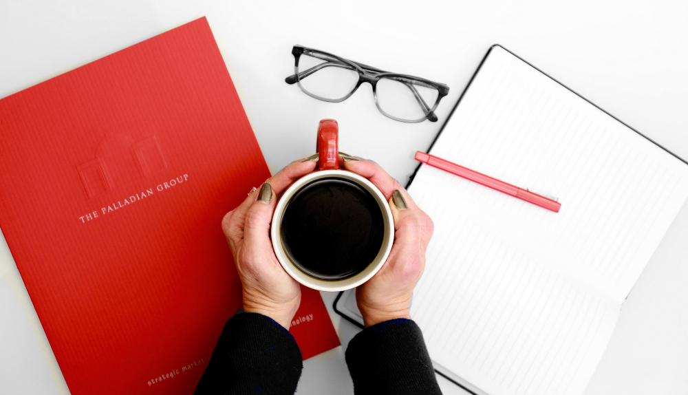 A red Palladian Group strategy booklet beside a notebook, glasses and a cup of coffee
