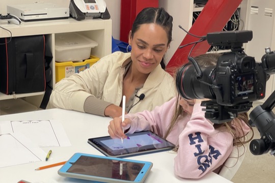 A woman and child working at a table while a camera records, in the firm’s Michelle Young Foundation project photograph