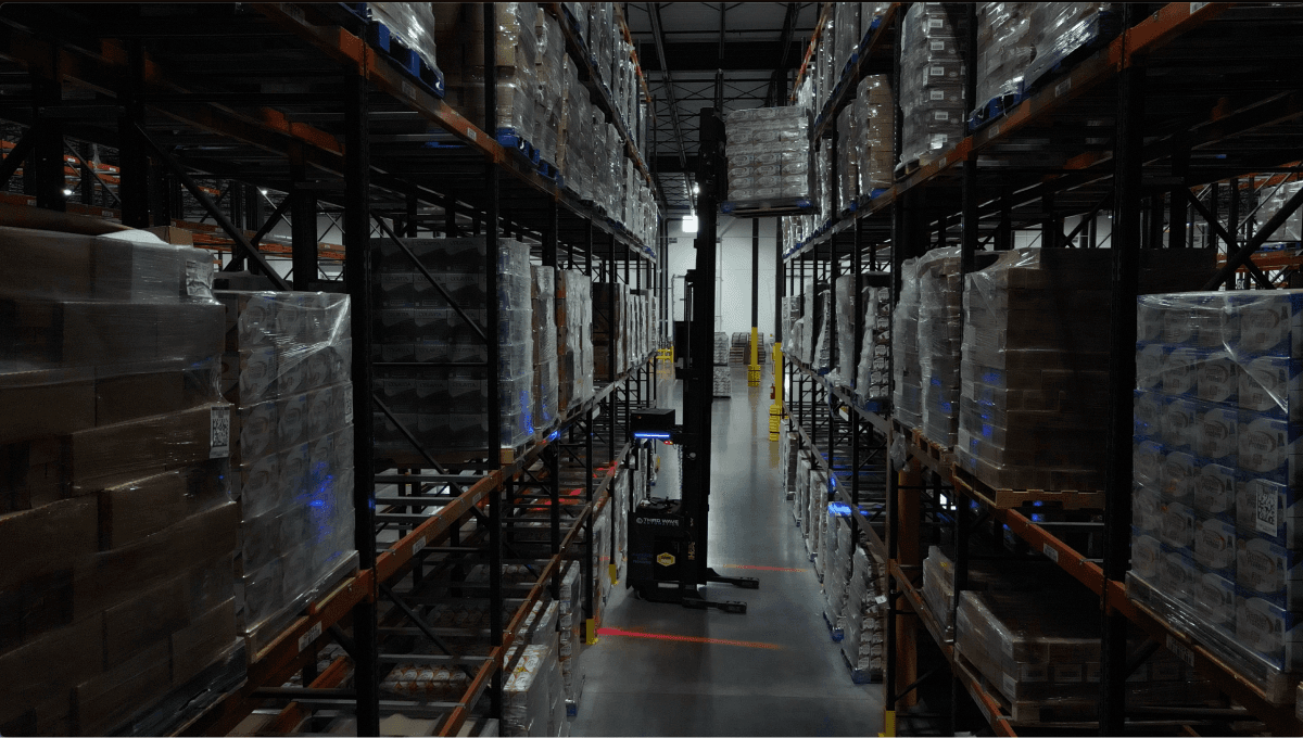 A Third Wave Automation autonomous reach truck working a dark warehouse aisle stacked with pallets