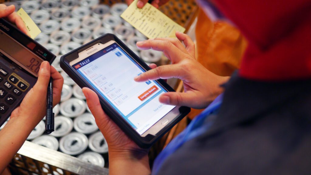 A worker enters production data into Tally on a tablet beside a digital scale and cans.