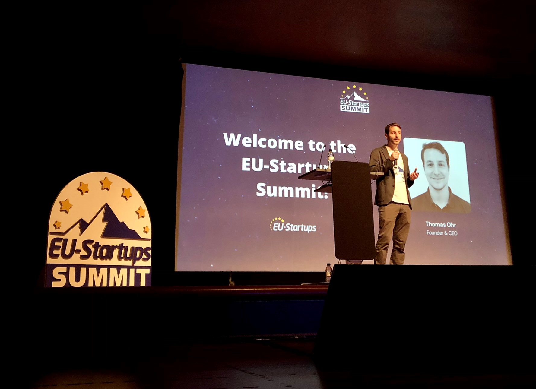 Thomas Ohr holding a microphone at the EU-Startups Summit in Barcelona, with the event welcome screen behind him