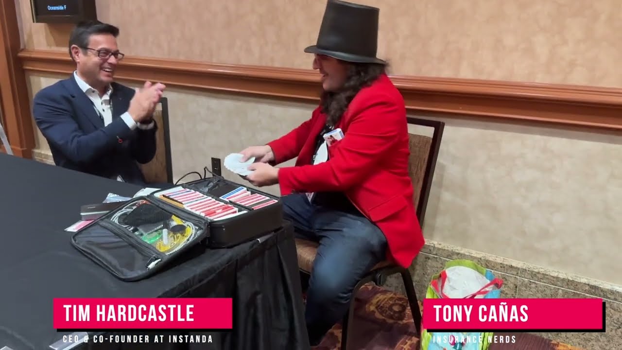 Tim Hardcastle laughing at a table opposite Tony Cañas, who wears a top hat and red jacket