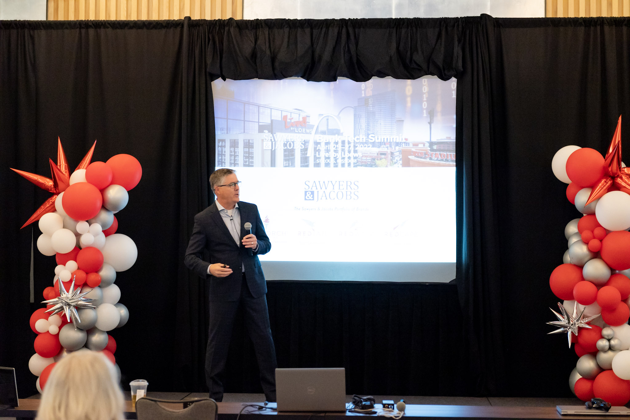 Tim Pannell speaking with a microphone at the 2022 Bank Tech Summit, between red and white balloon arrangements