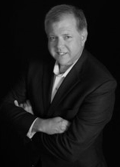 Official portrait of Tim Willis in a dark suit, photographed against a black background
