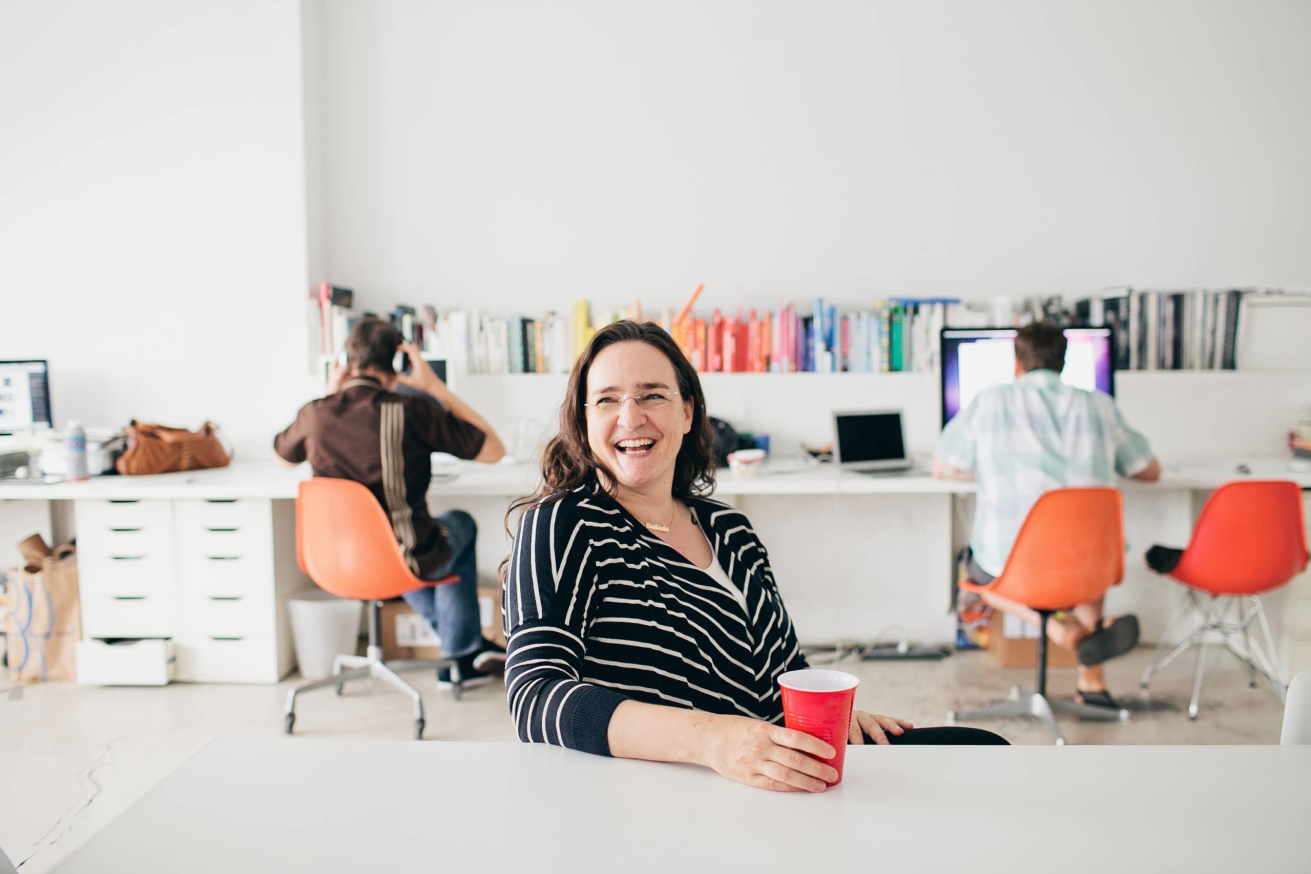 Tina Roth Eisenberg laughing at a table in a bright shared studio while coworkers sit behind her
