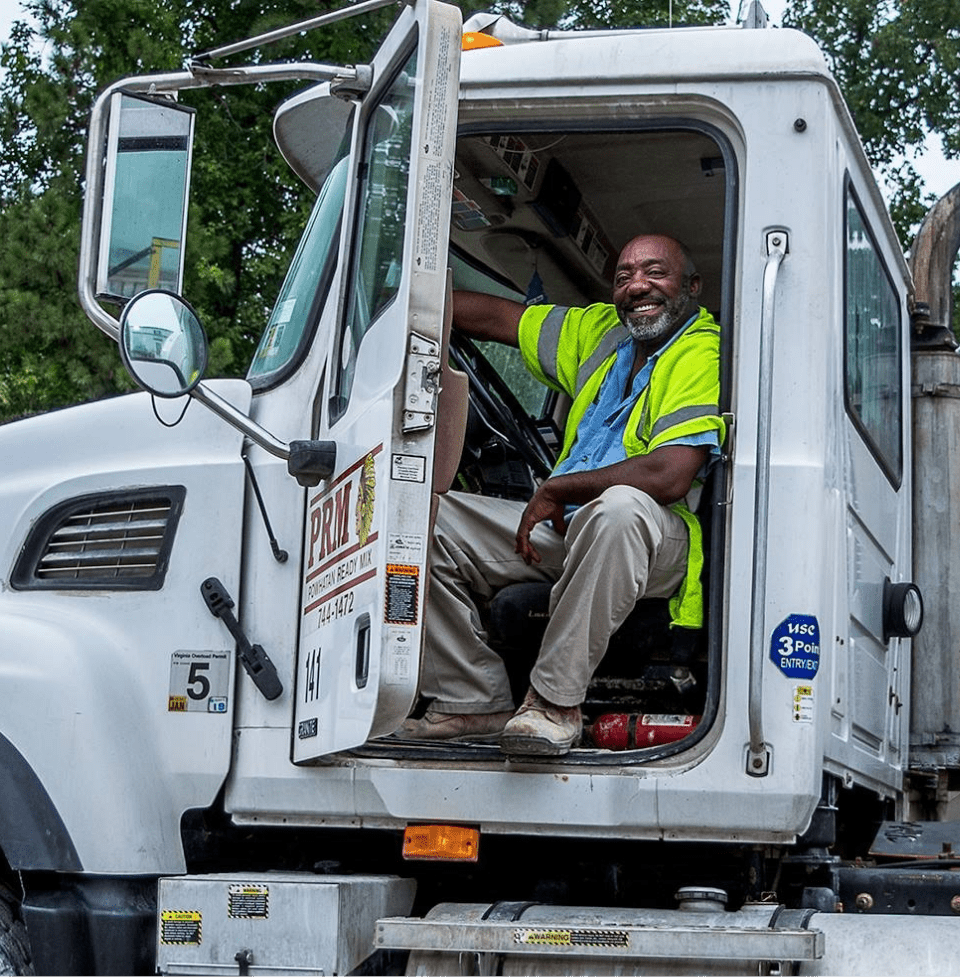 A smiling ready-mix driver seated in the open cab of a concrete truck