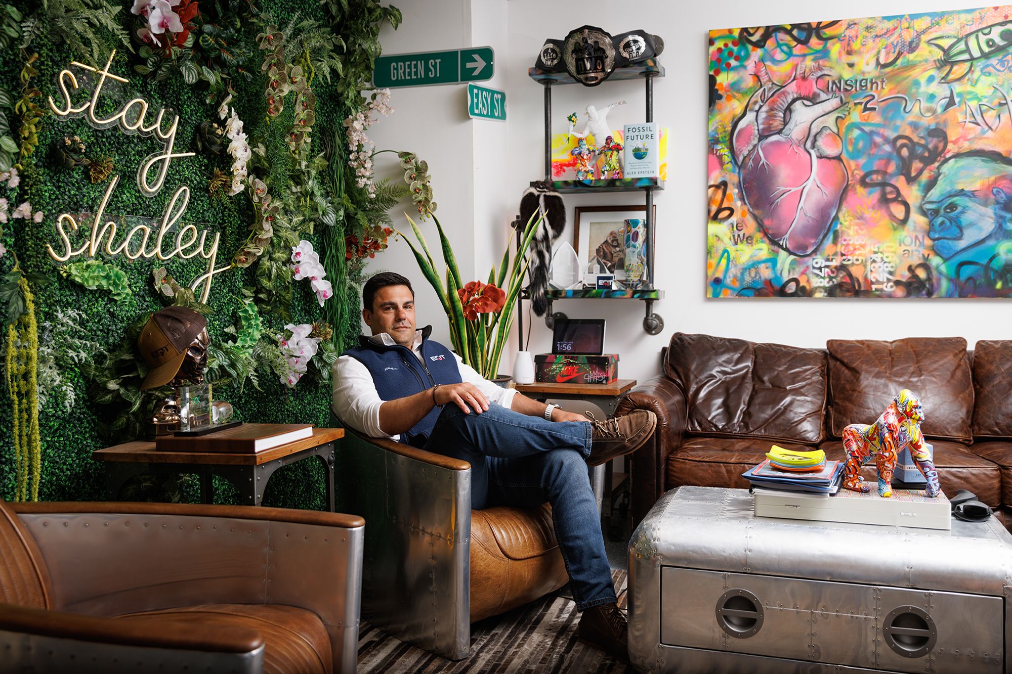 Toby Rice seated in a colorful room beside a plant wall reading Stay Shaley