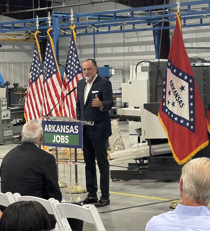 Todd Denton speaking at a lectern on Conveyor Technology’s factory floor, with manufacturing equipment and Arkansas flags behind him.