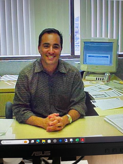 An archival photograph of Todd Greenbaum seated at a paper-covered office desk beside a CRT monitor