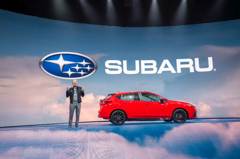 Tom Doll gestures on a Subaru presentation stage beside a red hatchback