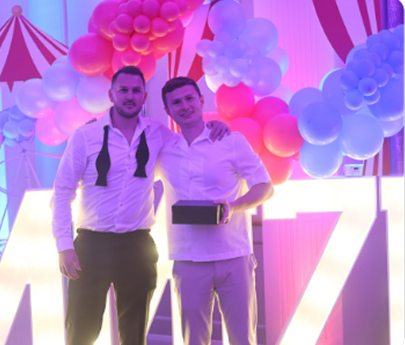 Tom Dunlop on the left, wearing an untied bow tie, poses beside another attendee against a colourful balloon backdrop.