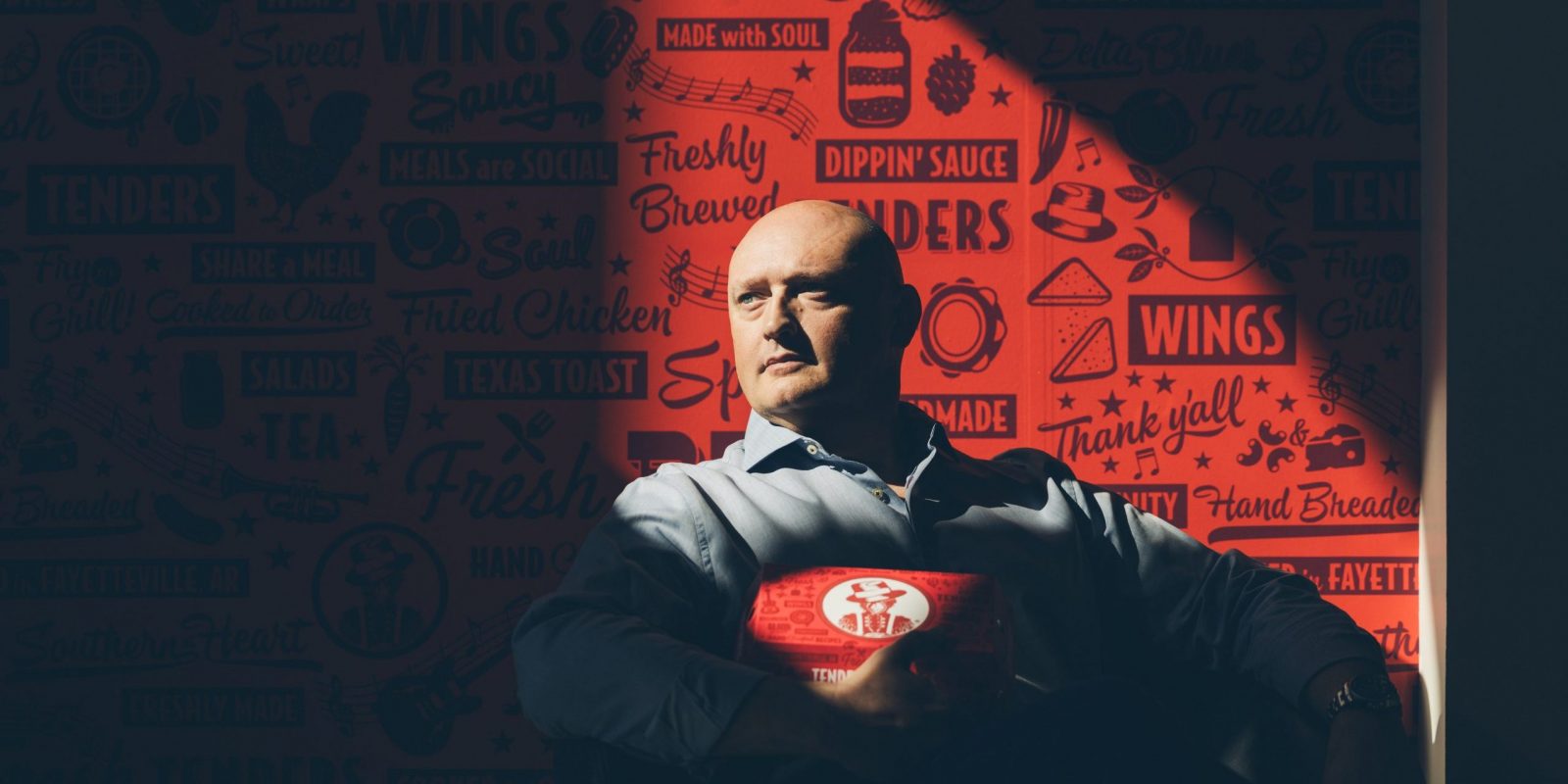 Tom Gordon seated in a Slim Chickens restaurant, holding a takeout box against a red illustrated wall.