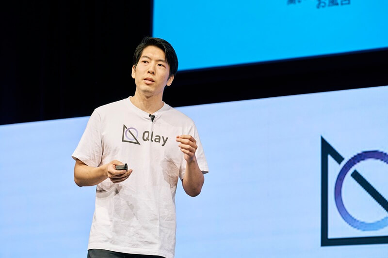 Tom Nakata presenting on stage in a Qlay t-shirt