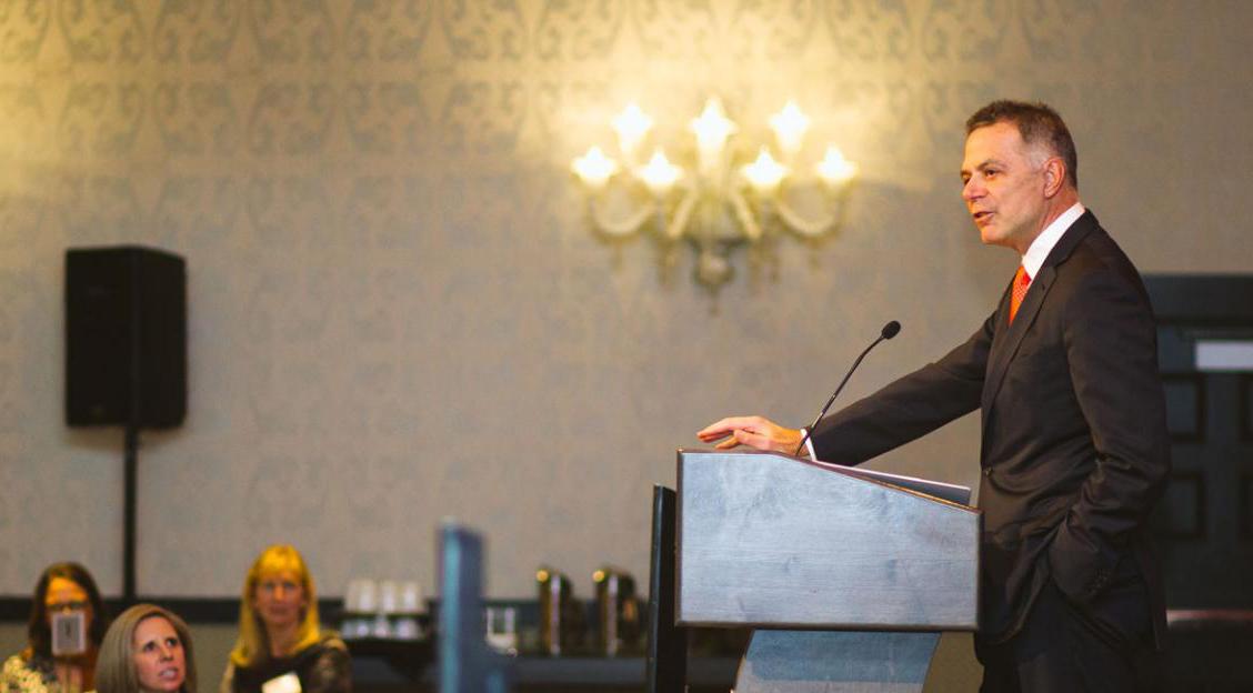 Tom Toomey speaking at a lectern, wearing a dark suit and orange tie