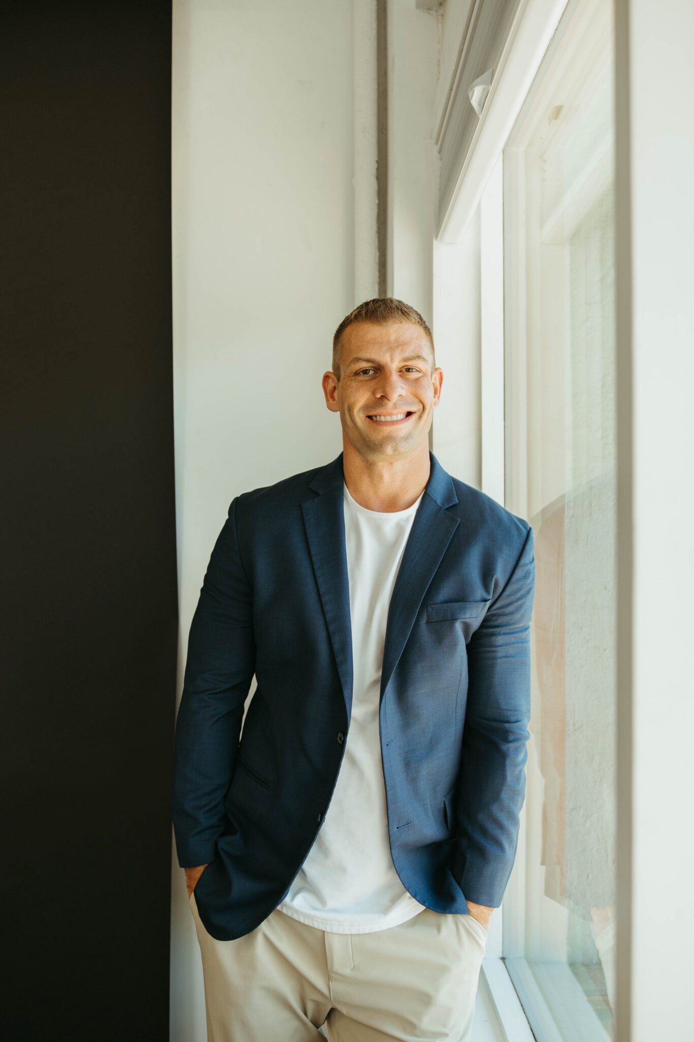 Tommy Mello standing beside a tall window in a blue jacket