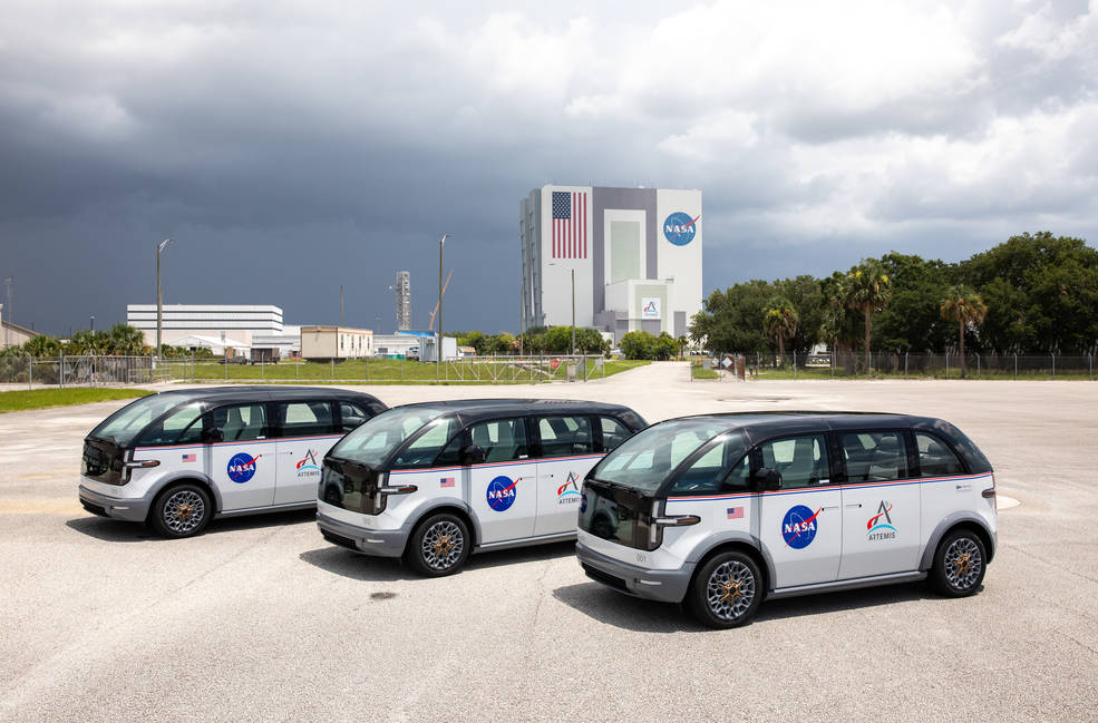 Three white Canoo crew transport vans parked in front of NASA’s Vehicle Assembly Building