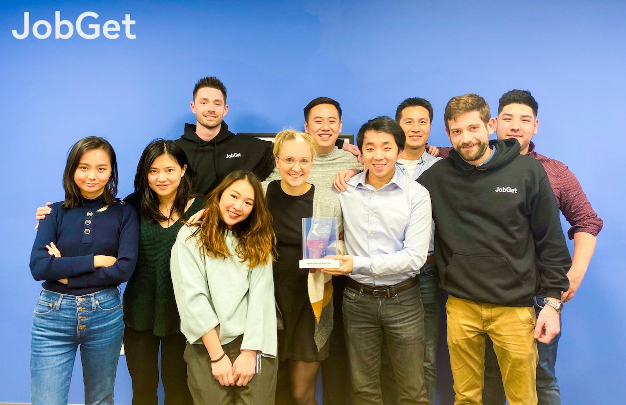 The early JobGet team gathered in front of a blue wall, holding an award