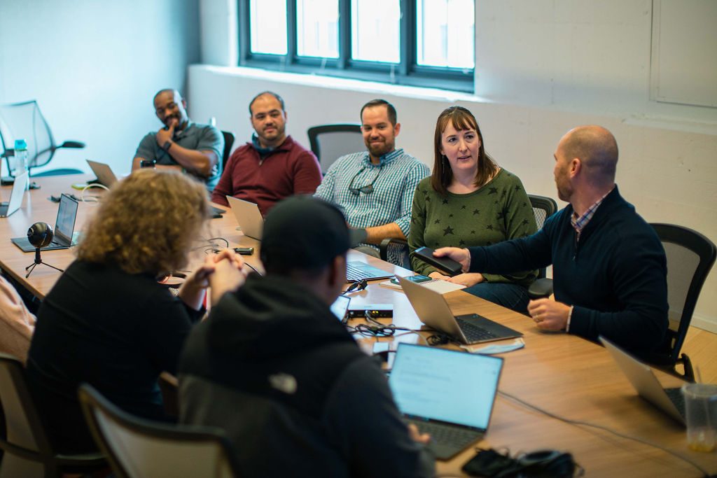 Colleagues in a TopBloc office photo sit around a long table with laptops.
