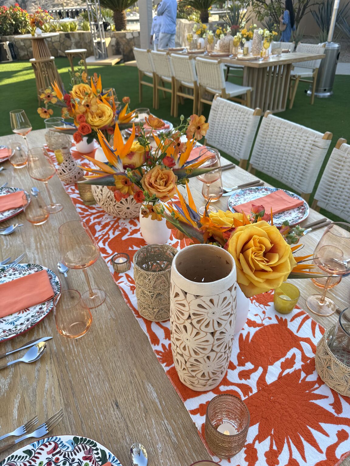 A long outdoor dining table with orange and yellow flowers, patterned plates and coral napkins at an Alkami President's Club event