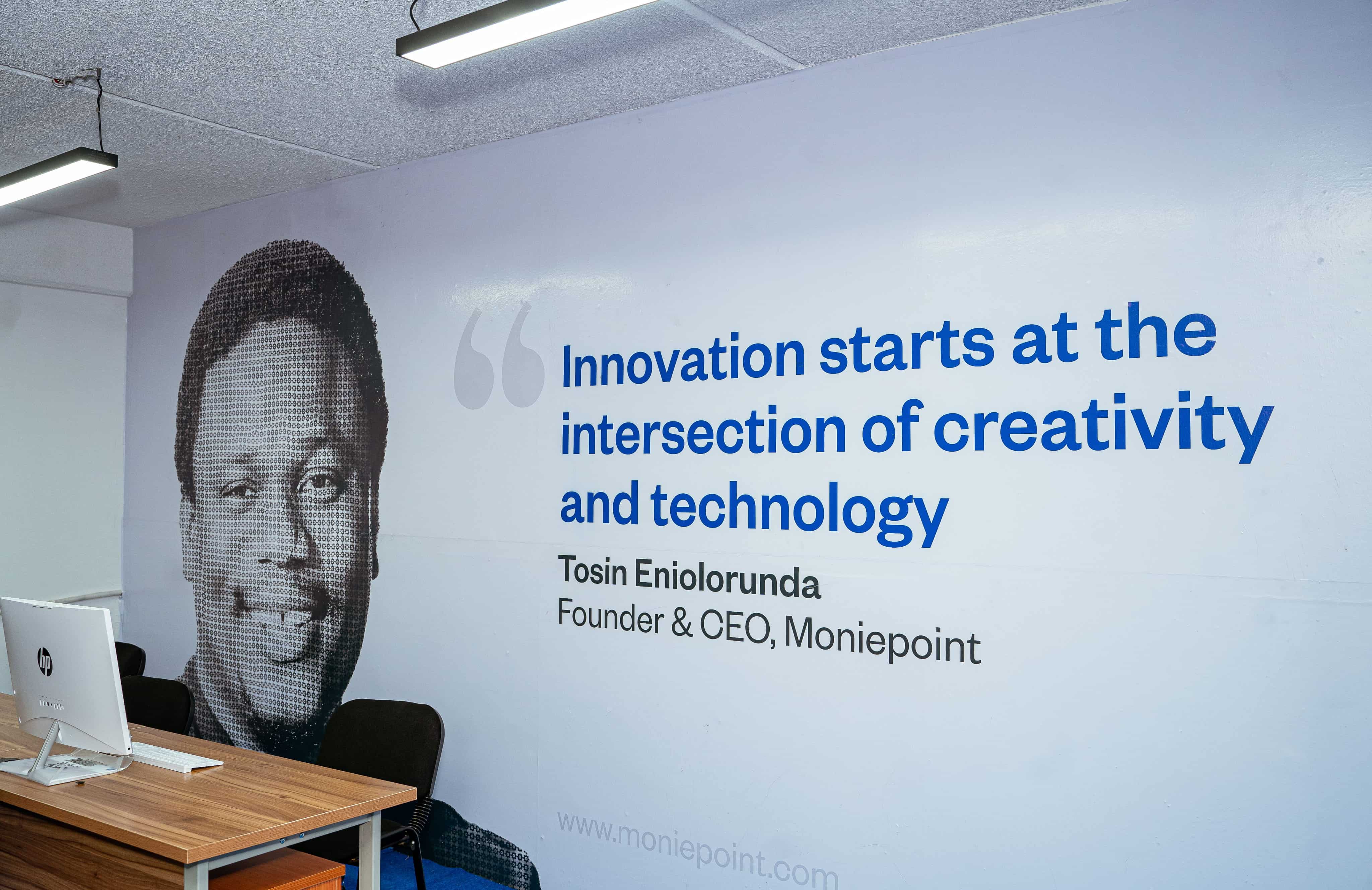 A computer workspace with a wall mural of Eniolorunda and a message about creativity and technology