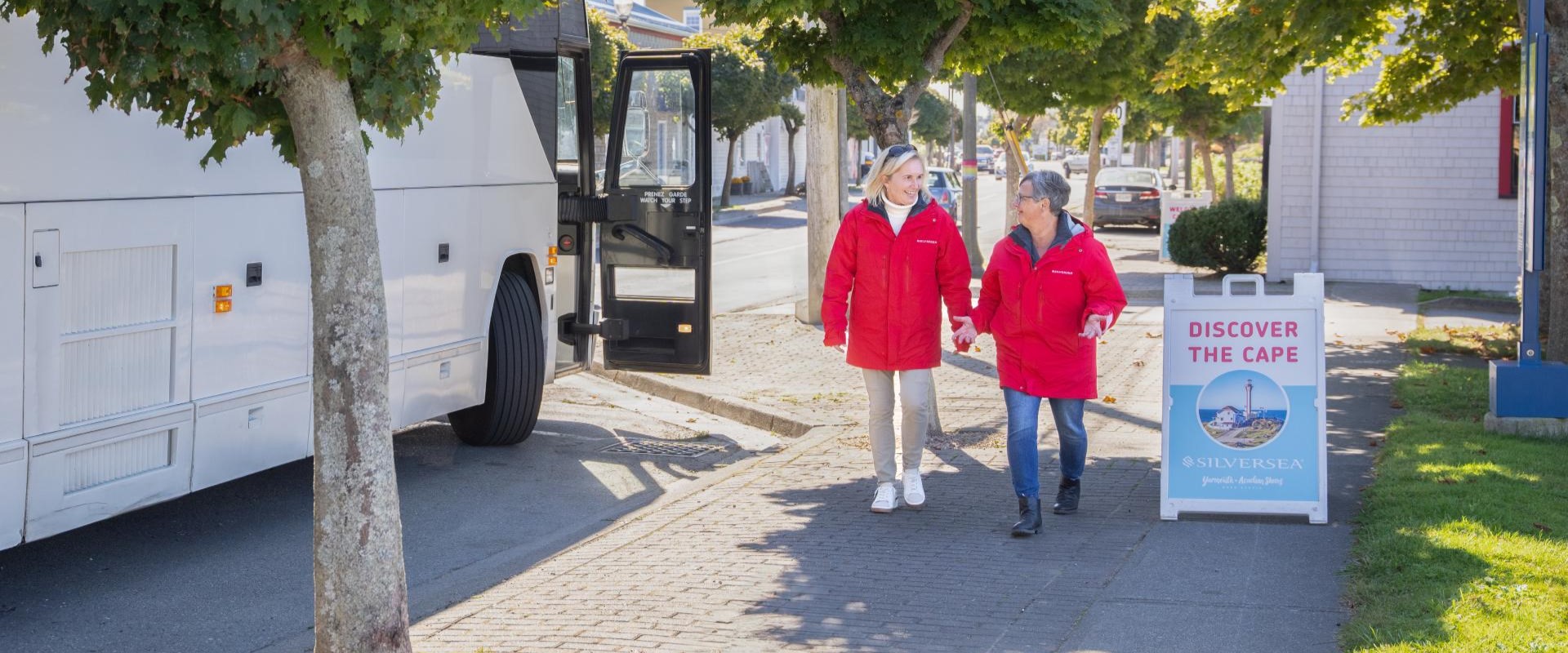 Two visitor hosts in red jackets walking beside a tour coach and a Discover the Cape sign in Yarmouth, Nova Scotia