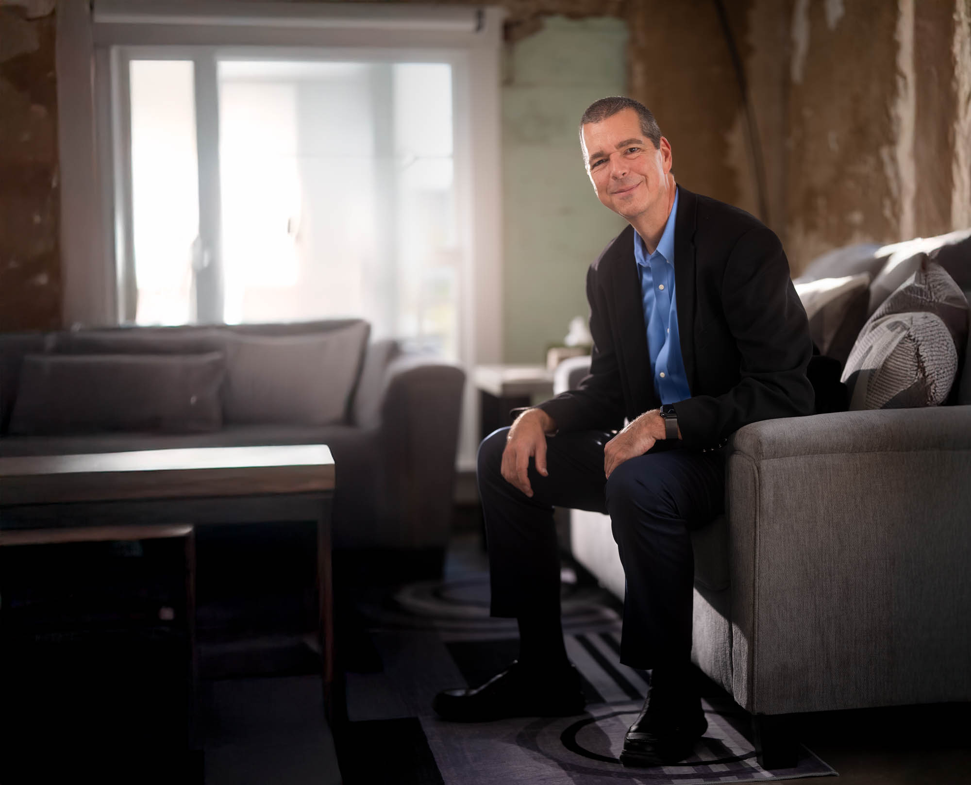 Travis Priest seated on a sofa in an interior with exposed brick and large windows
