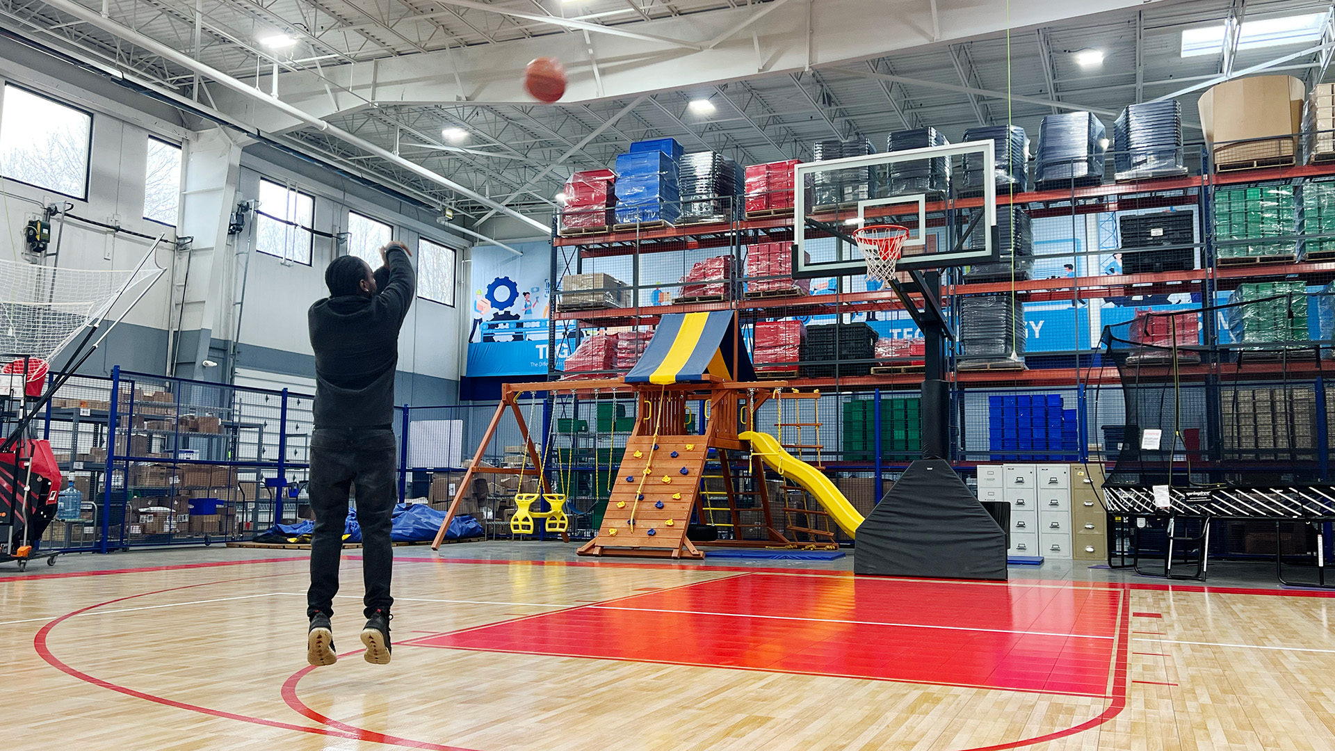 A person shoots a basketball on TRG’s indoor court, beside warehouse shelving and a children’s play structure.