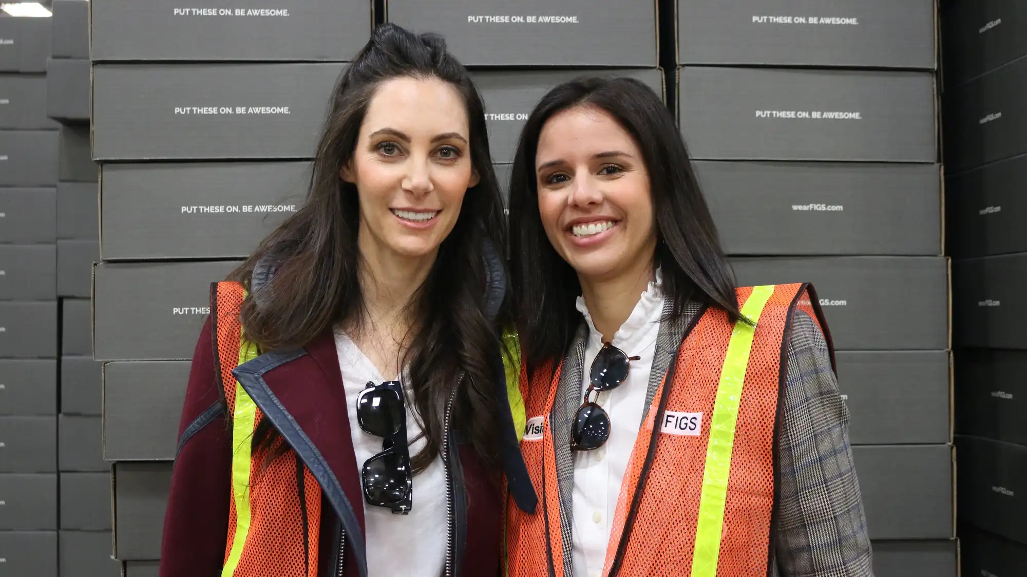 Heather Hasson and Trina Spear wearing safety vests in front of stacks of FIGS boxes