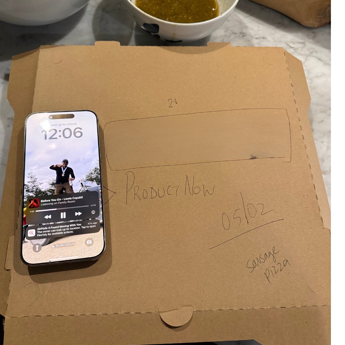 A pizza box bearing ProductNow handwriting beside a phone showing 12:06