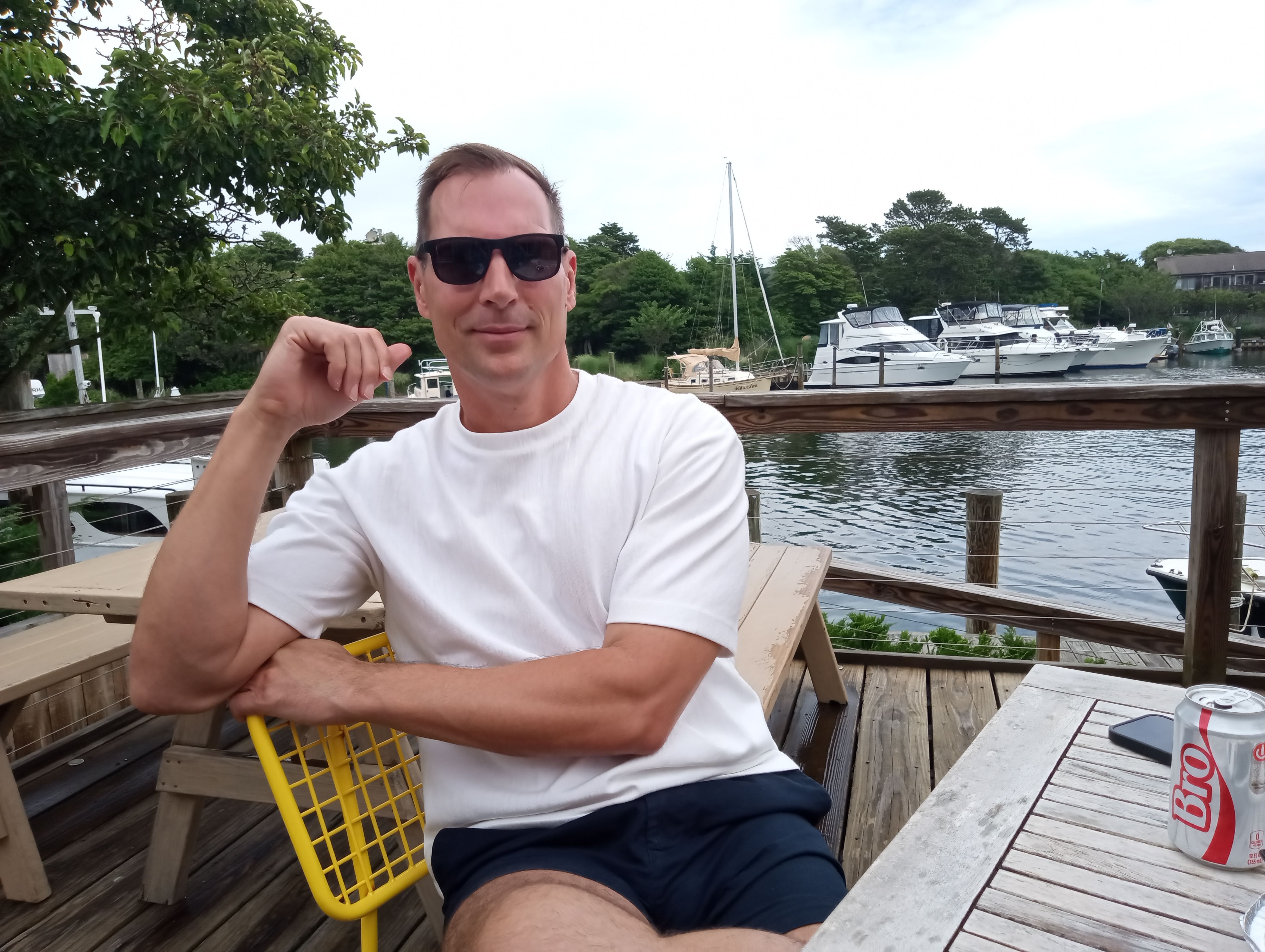 Tristan Schukraft in sunglasses and a white shirt, seated beside Fire Island Pines Harbor at The Canteen.