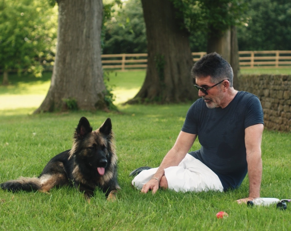 Simon Cowell sitting on the grass beside a German shepherd