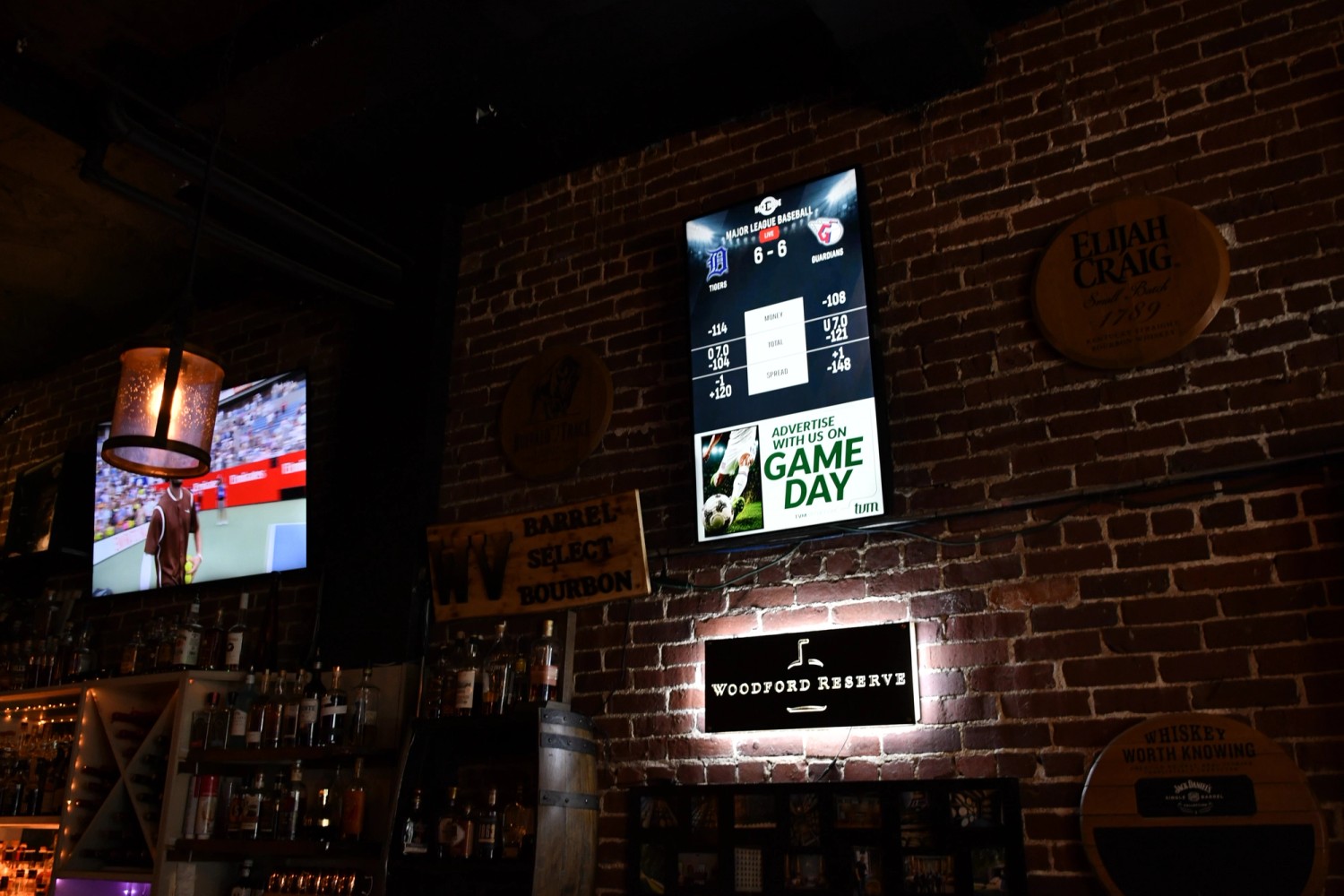A vertical TVM screen displaying baseball scores and odds beside a television in a brick-walled bar
